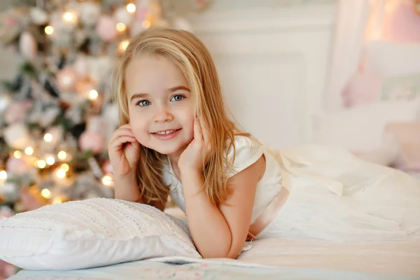 Muito bonito menina loira em um vestido branco segurando uma caixas de presente em um fundo de árvores de Natal no interior da casa — Fotografia de Stock