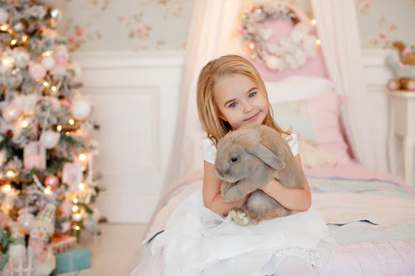 Muy linda niña rubia en un vestido blanco sosteniendo una caja de regalo sobre un fondo de árboles de Navidad en el interior de la casa — Foto de Stock