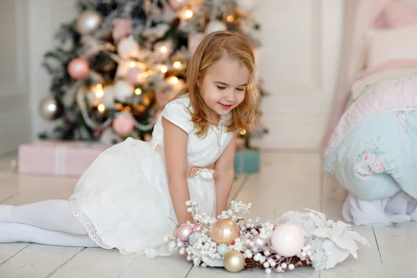 Muy linda niña rubia en un vestido blanco sosteniendo una caja de regalo sobre un fondo de árboles de Navidad en el interior de la casa — Foto de Stock