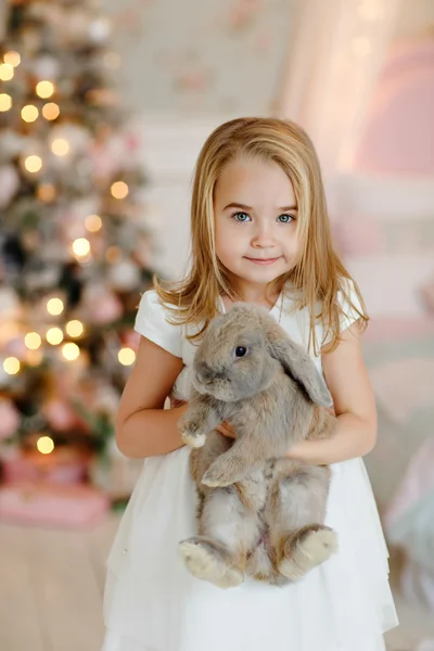 Velmi roztomilá malá holka blondýnka v bílých šatech drží dar krabice na pozadí vánočních stromků v interiéru domu — Stock fotografie