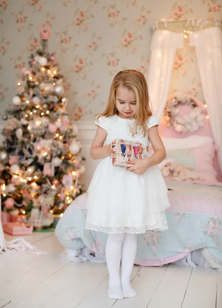 Muy linda niña rubia en un vestido blanco sosteniendo una caja de regalo sobre un fondo de árboles de Navidad en el interior de la casa — Foto de Stock