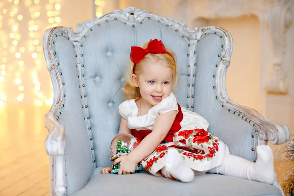 Babymeisje blonde in een rode jurk charmante vormt een gezicht, zitten — Stockfoto