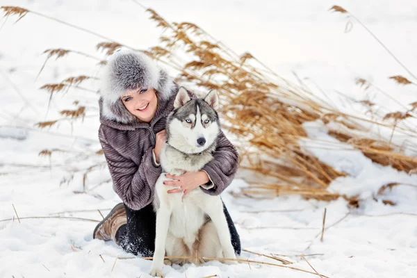 A menina no casaco cinza sorrindo ao lado de um cão husky cinza no fundo de inverno juncos amarelos — Fotografia de Stock