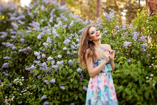 Hermosa chica glamour con el pelo largo y ondulado y hermoso maquillaje, en un vestido brillante se encuentra cerca de un Bush de lilas en verano y sonrisas saltando — Foto de Stock