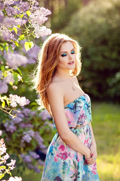 Красивая гламурная девушка с длинными волнистыми волосами и красивым макияжем, в ярком платье стоит рядом с Бушем сирени летом с закрытыми глазами — стоковое фото
