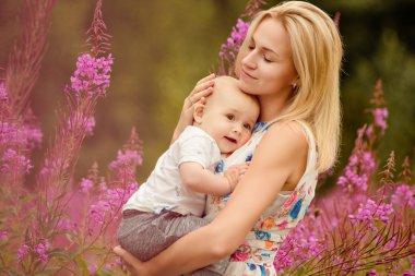 Güzel ince sarışın anne çok güzel bebek boy gülümseyerek çim fireweed arka plan üzerinde yaz aylarında hugs