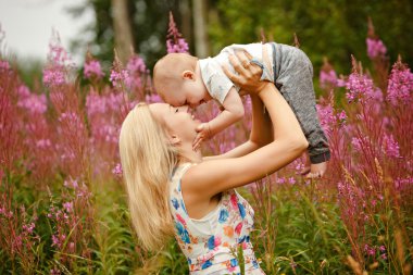 Güzel ince sarışın anne atar ve sevimli bebek boy gülümseyerek çim fireweed arka plan üzerinde yaz aylarında öper.
