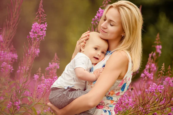 Hermosa delgado rubia mamá abraza adorable sonriente bebé niño en el fondo de hierba fireweed en el verano — Foto de Stock