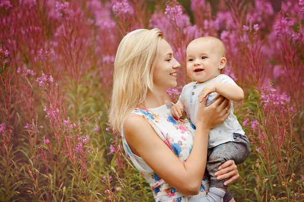 Schöne schlanke blonde Mutter umarmt entzückende lächelnde Baby Junge auf dem Hintergrund von Gras Feuerkraut im Sommer — Stockfoto