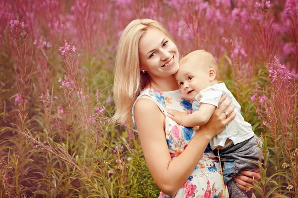 Krásná štíhlá blondýna matka objetí roztomilý usměvavý chlapeček na pozadí fireweed trávy v létě — Stock fotografie