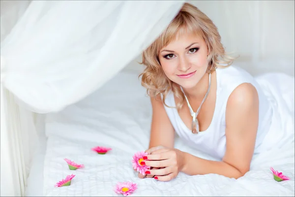 Roztomilá blondýnka na bílé posteli, obklopen malými květy — Stock fotografie