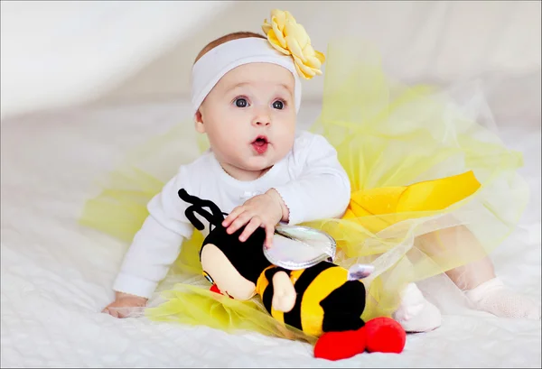 Uma criança pequena em uma saia amarela senta-se na cama com uma abelha de brinquedo — Fotografia de Stock