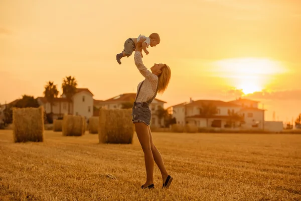 Sottile bella mamma bionda in jeans vestito bambino vomita sullo sfondo di campi di paglia al tramonto e sorride — Foto Stock