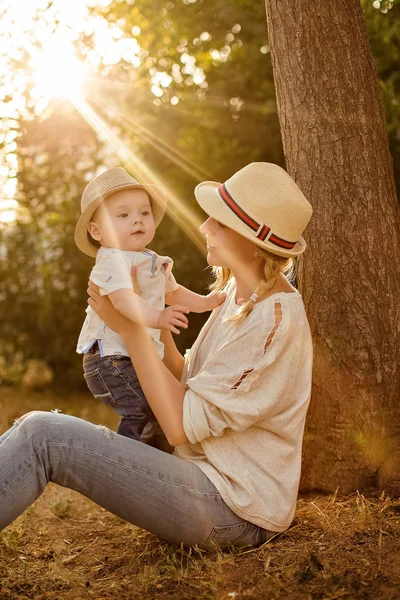 Magro bela mãe loira em jeans, uma camisa bege e um chapéu segurando um menino no fundo do parque ao pôr do sol e sorri — Fotografia de Stock