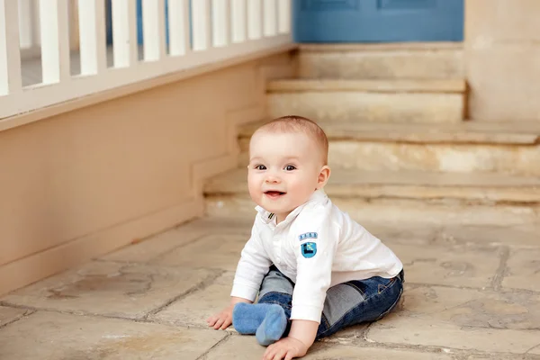 Niño muy lindo y de ojos grandes en jeans y una camiseta blanca sentada sobre un fondo de escaleras beige y sonrisas — Foto de Stock