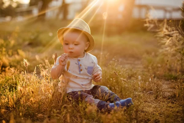 Pequeño sombrero encantador niño sentado en la hierba al atardecer en — Foto de Stock