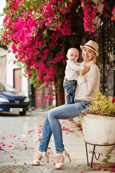 Giovane madre sorridente in un cappello di paglia che tiene in braccio un affascinante bambino in jeans in mezzo alla bouganville in fiore — Foto Stock