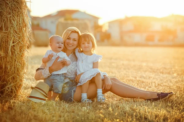 Una madre joven con dos hijos - un niño pequeño y una niña con un vestido blanco sentado sobre un fondo de campos de paja al atardecer y sonreír — Foto de Stock