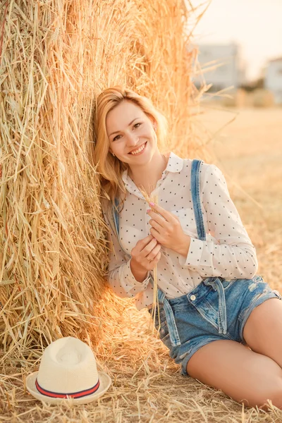 Чарівна красива дівчина блондинка в джинсовому костюмі сидить серед солом'яних полів на заході сонця і посміхається — стокове фото