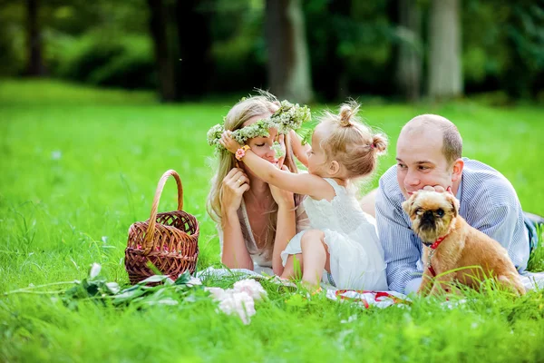 Mamá, papá, niña rubia y perro se acuestan juntos en la hierba y se prueban una corona —  Fotos de Stock