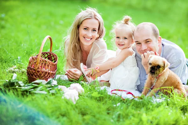 Mamá, papá, niña rubia y perro se acuestan juntos en la hierba y se ríen felizmente —  Fotos de Stock