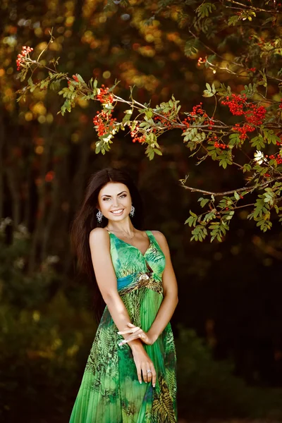 Retrato de uma sensual sexy muito bonita sorrindo menina morena com cabelos longos em um vestido verde na natureza sobre Rowan — Fotografia de Stock