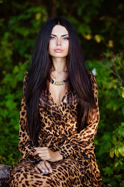 Portrait of a sexy sensual beautiful brunette girl with long hair in leopard yellow-black dress, sitting in the forest — Stock Fotó