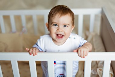 Little cute adorable little blond boy in a striped bodykit is in the nursery with white crib and laughs clipart