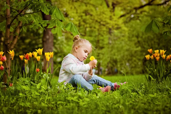 Very sweet beautiful blonde girl sits near the flower bed of red — Stock Photo, Image