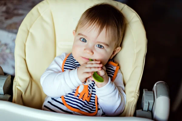 Little cute adorable toddler boy striped bib sits in a high chair and spoon nibbles — Stock Photo, Image