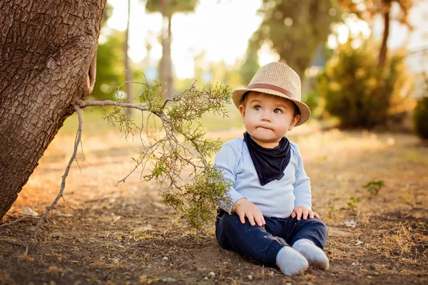 Kleine schattige babyjongen in een stro hoed en blauwe broek zitten met tuitte lippen, in de buurt van een boom op aarde bij zonsondergang in de zomer — Stockfoto