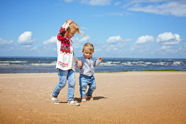 Sorella maggiore e fratello minore Bionde camminano lungo la spiaggia, tenendosi per mano, contro il mare . — Foto Stock