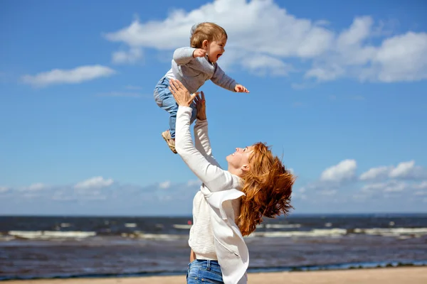 Pelirroja mamá en jeans y un suéter blanco arroja a su hijo joven contra el mar y las nubes —  Fotos de Stock