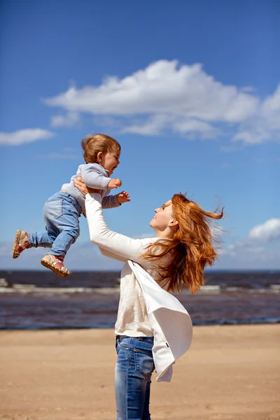 Pelirroja mamá en jeans y un suéter blanco arroja a su hijo joven contra el mar y las nubes —  Fotos de Stock