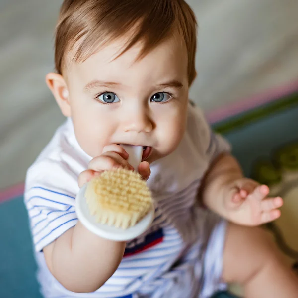 Zeer schattige kleine blonde jongen kauwt op de borstel voor het haar, zijn tanden, zittend op het gekleurde tapijt krassen — Stockfoto