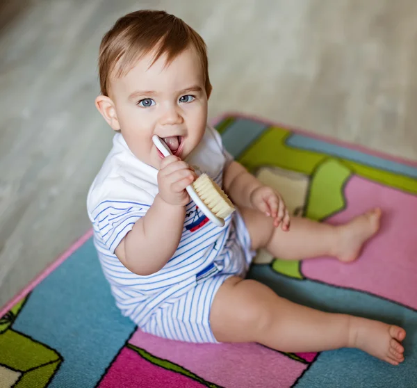 Très mignon petit garçon blond mâche sur la brosse pour les cheveux, se gratter les dents, assis sur le tapis coloré — Photo