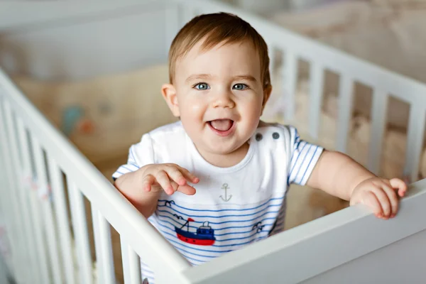 Pequeno menino louro adorável bonito em um kit de corpo listrado está no berçário com berço branco e ri — Fotografia de Stock