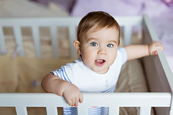 Pequeno menino louro adorável bonito em um kit de corpo listrado está no berçário com berço branco e ri — Fotografia de Stock