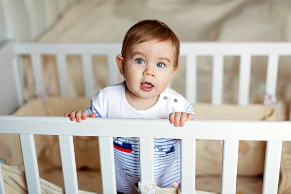 Little cute adorable little blond boy in a striped bodykit is in  the nursery with white crib and holds the handle