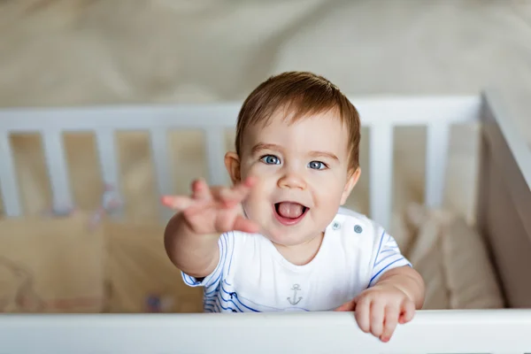 Little cute adorable little blond boy in a striped bodykit is in  the nursery with white crib and holds the handle — Stock Photo, Image