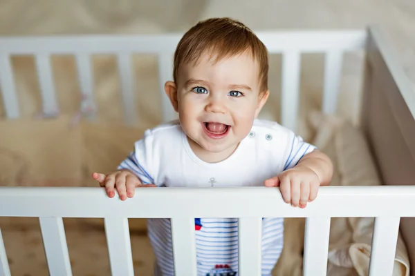 Poco lindo adorable niño rubio en un bodykit rayas está en el vivero con cuna blanca y se ríe — Foto de Stock