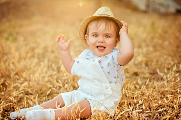 Pequeno menino gordinho adorável em um chapéu de palha e um terno branco sentado em um campo com espigas de trigo ao pôr do sol no verão — Fotografia de Stock