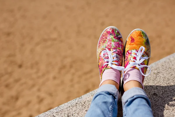 Gambe femminili in jeans e una brillante sneakers floreali sullo sfondo della sabbia in estate — Foto Stock