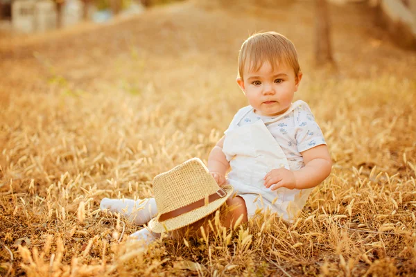 Piccolo affascinante bambino paffuto in una tuta bianca con un cappello, seduto in un campo nelle spighette nei caldi raggi del sole al tramonto in estate — Foto Stock