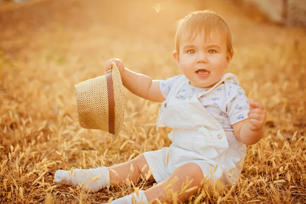 Pequeño niño regordete encantador en un traje blanco sosteniendo un sombrero, sentado en un campo en las espiguillas en los cálidos rayos del sol poniente en el verano —  Fotos de Stock