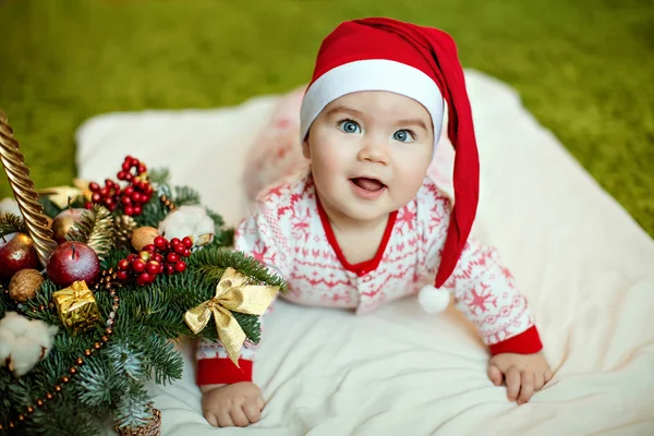 Pequeno menino encantador em chapéus vermelhos de Papai Noel e pijama com flocos de neve sorrindo, deitado perto de uma pequena árvore de Natal com brinquedos — Fotografia de Stock