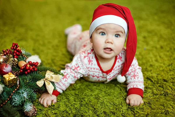 Pequeno menino encantador em chapéus vermelhos de Papai Noel e pijama com flocos de neve sorrindo, deitado perto de uma pequena árvore de Natal com brinquedos — Fotografia de Stock