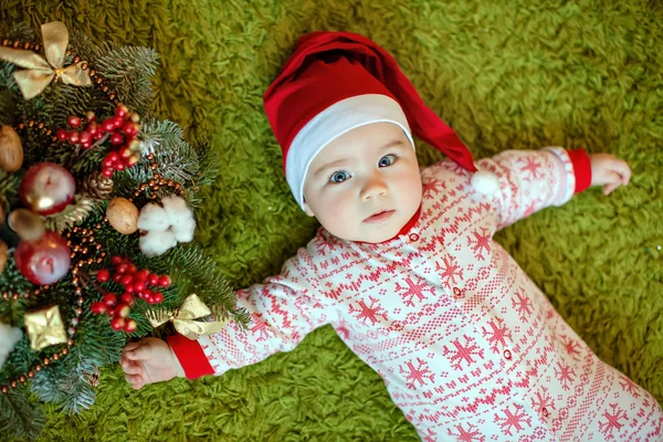 Pequeno menino encantador em chapéus vermelhos de Papai Noel e pijama com flocos de neve sorrindo, deitado perto de uma pequena árvore de Natal com brinquedos — Fotografia de Stock