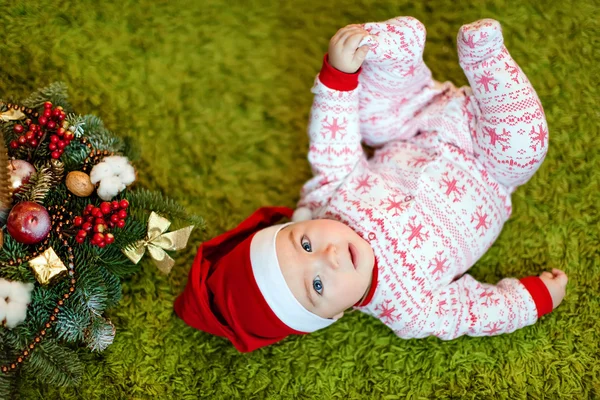 Pequeno menino encantador em chapéus vermelhos de Papai Noel e pijama com flocos de neve sorrindo, deitado perto de uma pequena árvore de Natal com brinquedos — Fotografia de Stock