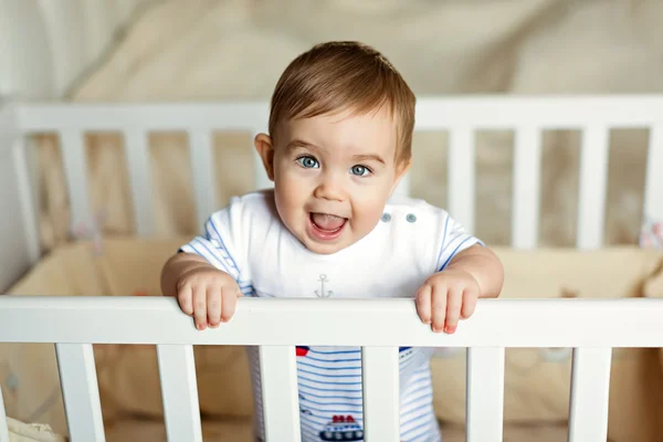 Poco lindo adorable niño rubio en un bodykit rayas está en el vivero con cuna blanca y se ríe Imágenes De Stock Sin Royalties Gratis